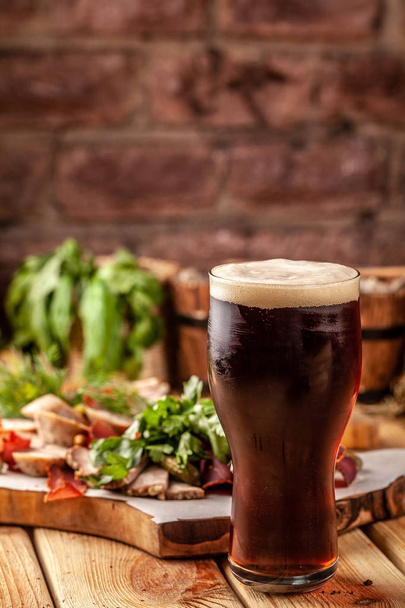 Concept of Georgian restaurant. A glass of black beer with foam stands on a wooden table, next to it is a beer snack, dried sausage, basturma, smoked meat and parsley with dill. - Photo, image