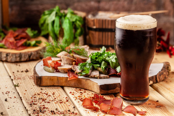 Concept of Georgian restaurant. A glass of black beer with foam stands on a wooden table, next to it is a beer snack, dried sausage, basturma, smoked meat and parsley with dill. - Photo, image