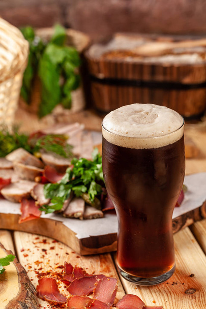 Concept of Georgian restaurant. A glass of black beer with foam stands on a wooden table, next to it is a beer snack, dried sausage, basturma, smoked meat and parsley with dill. - 写真・画像