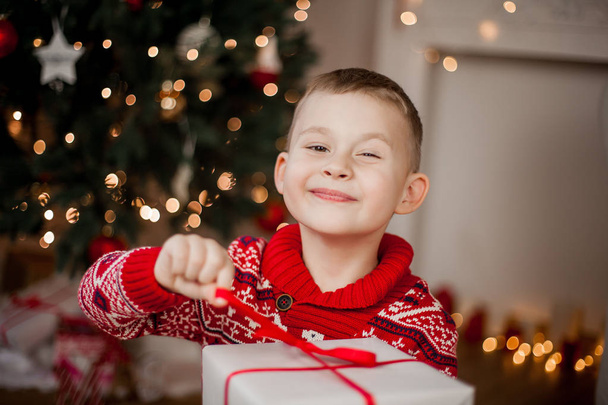 Pieni söpö poika punaisessa joulupuserossa istuu joulukuusen lähellä. Iloisen 4-vuotiaan pojan muotokuva lahjapakkauksella. Juhlapyhät. Joulu. Uusi vuosi
. - Valokuva, kuva