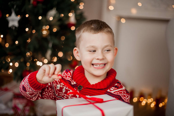 A piros karácsonyi pulóvert aranyos kisfiú ül a karácsonyfa közelében. Portréja egy boldog fiú 4 éves díszdobozban. Ünnepek. Karácsonyi. Új év. - Fotó, kép