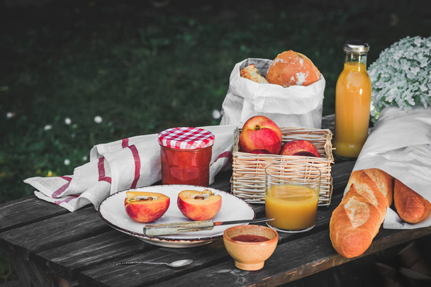 Assortment of food for picnic on wooden table. Plate with apricots, little jar with jam , variety of bread, glace of juice. Cozy summer breakfast - Photo, image