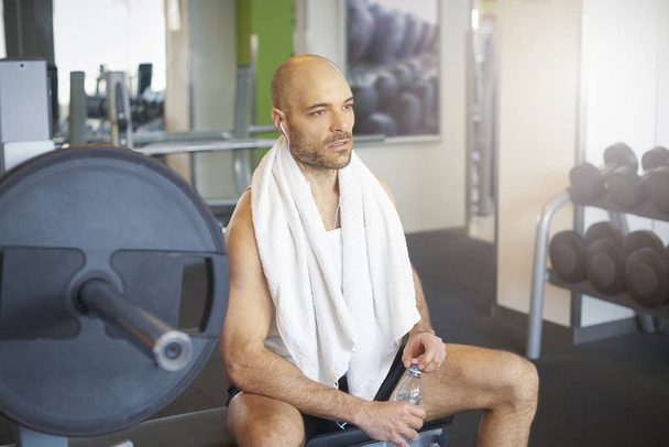 Shot of sporty man sitting in gym after workout and drinking a bottle of water. - 写真・画像