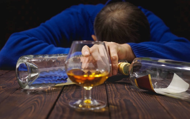 Male alcoholic with glass of whiskey and bottle sleeping on table at night. Alcohol and addiction. Concep - Foto, Imagen