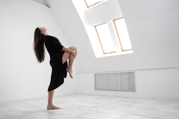 A young female dancer in a black dress is stretching by the window. White Hall, the working class, yoga asanas. - Fotoğraf, Görsel