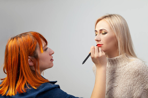 Make-up artist dívka s červenými vlasy dává červenou rtěnkou na rtech blondýna model sedí se zavřenýma očima v ateliéru na bílém pozadí s make-up víčka v orientálním stylu. - Fotografie, Obrázek