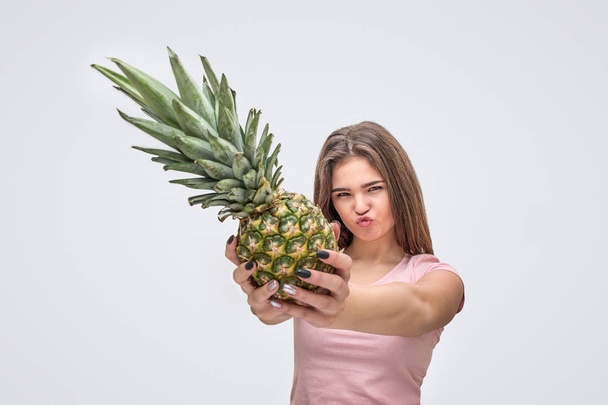 Serious young woman show pineapple from hands. She look on camera. Isolated on grey background. - Fotoğraf, Görsel