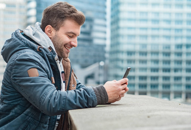Счастливый молодой человек печатает на телефоне, на улице. Вид сбоку или профиль ухмылочного парня, держащего смартфон
 - Фото, изображение