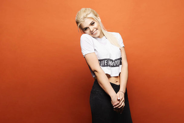 Fashionable, happy and smiling blonde model girl with perfect sexy body and with bright makeup in stylish black jeans and in white t-shirt posing at orange background, isolated - Φωτογραφία, εικόνα