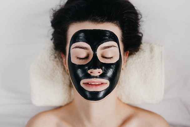 Beautiful young girl with eyes closed relaxing with applied a black mask to her face. Woman with purifying black mask on her face. Skin care and beauty concept. - Foto, imagen