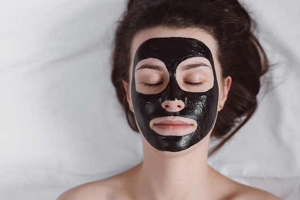 Beautiful young girl with eyes closed relaxing with applied a black mask to her face. Woman with purifying black mask on her face. Skin care and beauty concept. - Fotoğraf, Görsel