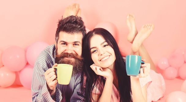 Man and woman on smiling faces lay, pink background. Couple in love drink coffee in bed. Couple relax in morning with coffee. Family tradition concept. Man and woman in domestic clothes, pajamas - Foto, immagini