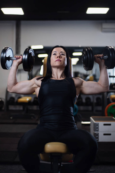 Athletic fitness girl trains with dumbbells in the gym. Pumps the deltoid muscles - Photo, Image