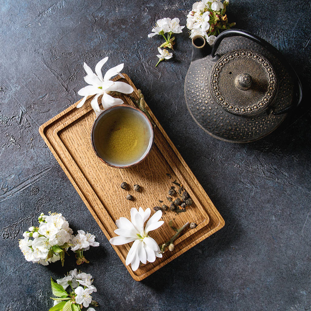 Traditional ceramic cup of hot green tea on wooden board with black iron teapot, spring flowers white magnolia, cherry blooming branches. Blue texture background. Top view, space. Square image - Foto, imagen