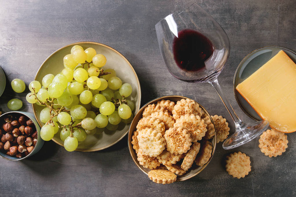 Wine snack. Cheese, grapes, nuts, cheese crackers cookies, honeycombs with laying glass of red wine and knife over dark texture background. Flat lay, space - Photo, image