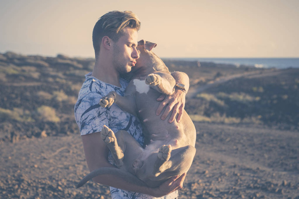 Young attractive blonde man model hug with love and friendship his own cute puppy dog amstal in outdoor leisure activity and scenic desertic place with ocean in background - Photo, Image