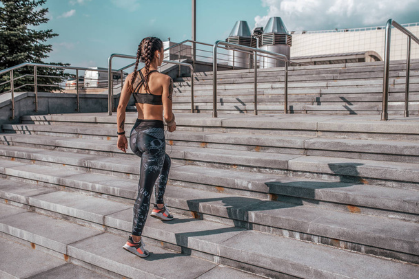 Girl in sportswear, view from rear. In his hand a smartphone listening to music with headphones. Rises on the stairs, in summer in city. Sports and healthy lifestyle. Free space for text. - Fotografie, Obrázek