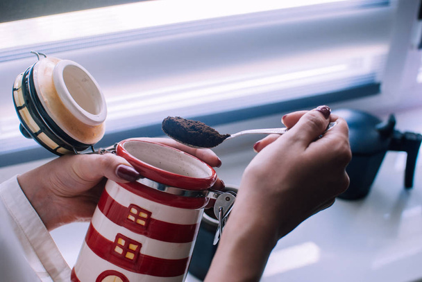 Una chica sosteniendo una cucharada de café llena una cafetera de géiser de metal negro. Luz brillante y soleada que entra por la ventana. Ambiente acogedor por la mañana. Manera perfecta de empezar un día con energía y alegría
. - Foto, Imagen