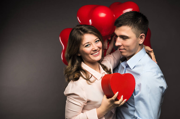 Νεαρό ζευγάρι με κόκκινα μπαλόνια κοντά γκρίζο τοίχο σε σχήμα καρδιάς - Φωτογραφία, εικόνα
