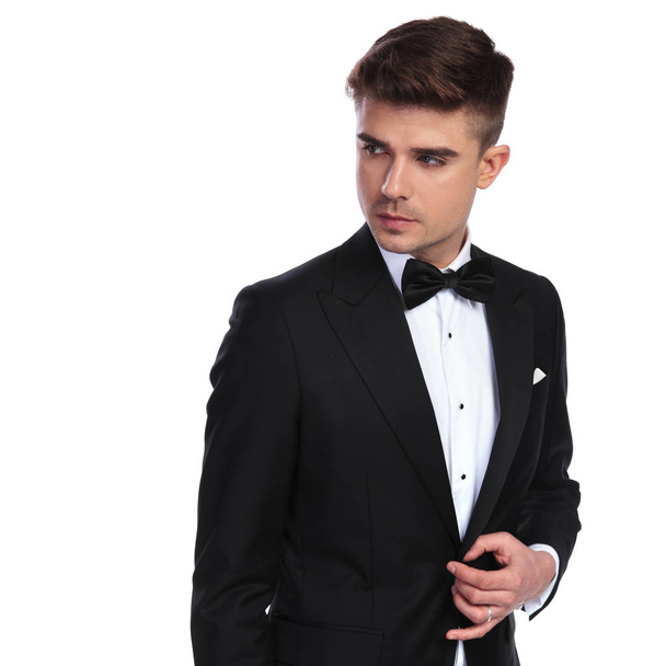 πορτρέτο του περίεργος επιχειρηματίας με παπιγιόν unbuttoning του με μαύρο κοστούμι, ενώ ψάχνει πλευρά και να στέκεται πάνω σε λευκό φόντο - Φωτογραφία, εικόνα