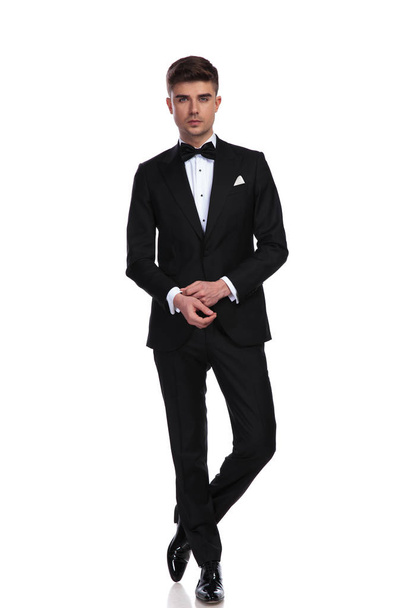 attractive groom fixing cuffs while standing on white background with legs crossed, full length picture - Foto, imagen