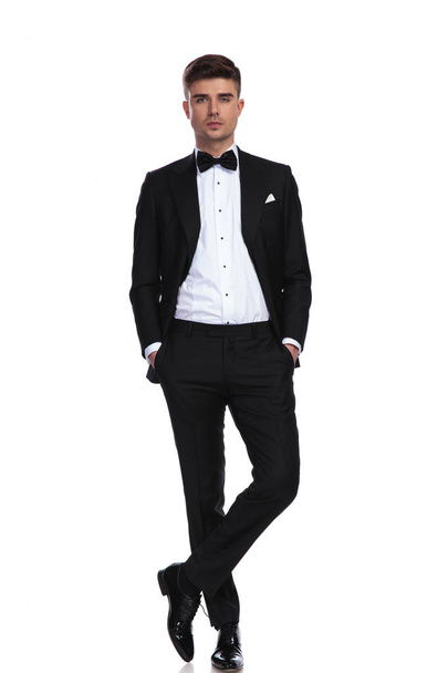 young groom in black tuxedo standing with hands in pockets on white background with legs crossed - Foto, Bild