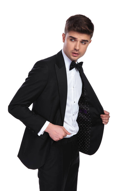 портрет привлекательного бизнесмена, держащего воротник черного костюма и смотрящего в сторону, стоя на белом фоне
 - Фото, изображение