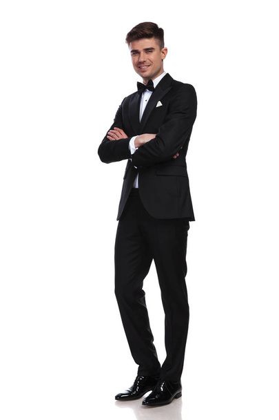 smiling groom in black tuxedo standing on white background with arms folded, full body picture - Фото, зображення