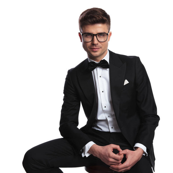 portrait of smiling nerd wearing a black tuxedo sitting on wooden chair on white background and holding palms together - Photo, Image