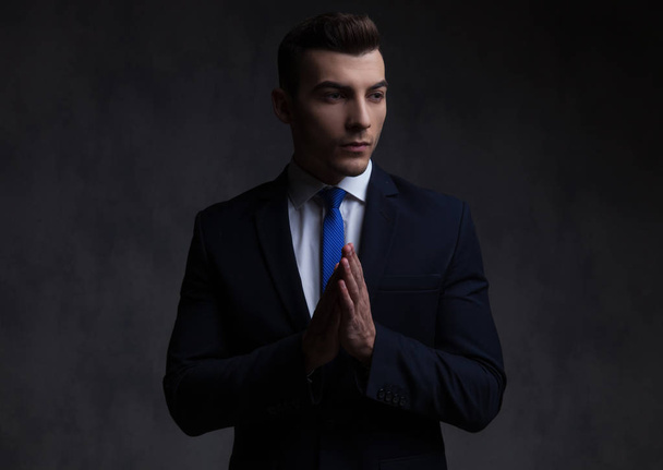 portrait of curious businessman praying and looking to side while standing on black background - Fotografie, Obrázek