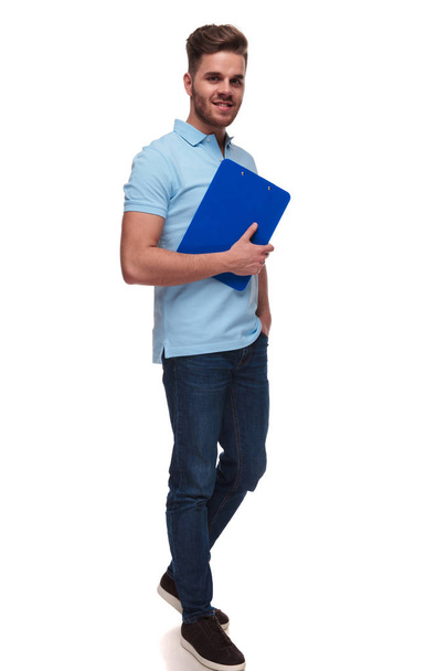 ελκυστικό casual άνδρα που φοράει κοντό μπλε polo κατέχει μπλε αρχεία και τα βήματα για να την πλευρά με τα χέρια στις τσέπες σε άσπρο φόντο, πλήρη εικόνα σώματος - Φωτογραφία, εικόνα