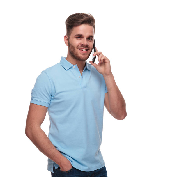 портрет расслабленного мужчины в рубашке для поло, говорящего по телефону, стоя на белом фоне с рукой в кармане
 - Фото, изображение