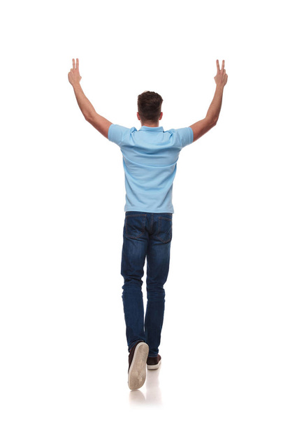 back view of casual man celebrating and making peace sign while stepping on white background, full length picture - Photo, Image