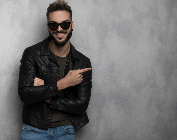 portrait of smiling casual man wearing leather jacket pointing to side while leaning against a grey wallpaper background - Photo, Image