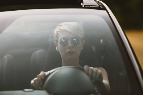 Молодая блондинка привлекательная женщина улыбается и смотрит прямо во время вождения автомобиля
. - Фото, изображение
