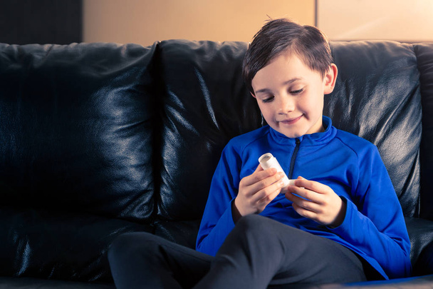 αγόρι 8 χρονών φορώντας αθλητική ενδυμασία κρατώντας μια συσκευή εισπνοών άσθμα που κάθεται στον καναπέ. Έννοιες: την υγεία, ασθένεια, ντόπινγκ - Φωτογραφία, εικόνα
