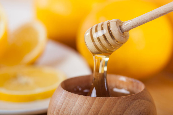 το μέλι σε ένα ξύλινο μπολ με κουτάλα μελιού και λεμονιού σε ένα ταμπλό με ξύλινη κουζίνα. - Φωτογραφία, εικόνα