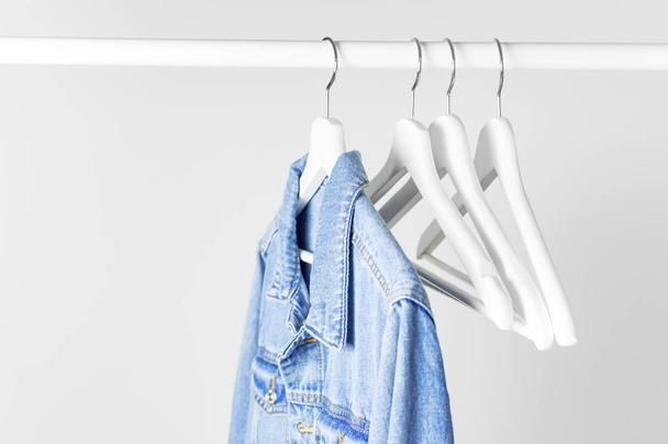Blue denim jacket on white wooden coat hanger on a rod against light gray wall flat lay copy space. Denim, fashionable jacket, women's or men's trend clothing, fashion background. Store concept, sale - Photo, image