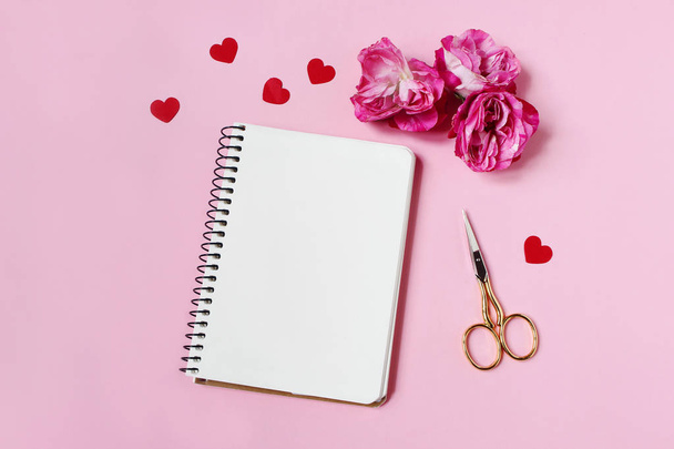 Wedding, birthday or Valentines stationery mockup scene. Pink roses, red heart shape paper confetti, blank notebook and golden scissors on pink background. Flat lay, top view. - Photo, image