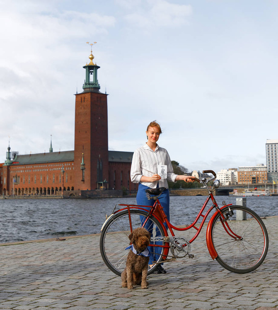 Sztokholm - 22 września 2018: Kobieta i pies noszący zabytkowy rower przed ratuszem w Sztokholmie podczas imprezy Bike in Tweed 22 września 2018 w Sztokholmie, Szwecja - Zdjęcie, obraz