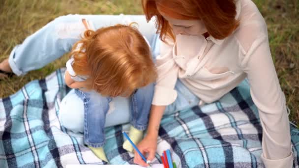 Vista superior de la niña pelirroja y su madre dibujando en el parque. Dulce hija se ve cómo su madre pinta con lápiz. Vista superior
 - Imágenes, Vídeo