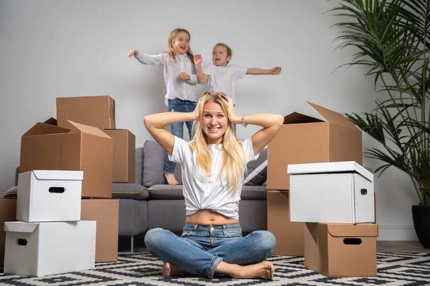 Photo of displeased blonde sitting on floor among cardboard boxes and boy, girl jumping on sofa - Photo, Image