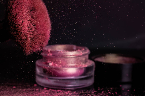 Sombras cor-de-rosa, brilho na escova cosmética e um frasco com uma tampa sobre um fundo escuro
. - Foto, Imagem