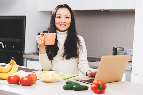 Picture of happy asian brunette with mug and laptop standing at table with vegetables and fruits - Photo, Image