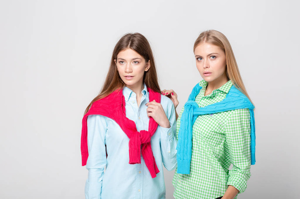 happy girlfriends women in shirts and a sweater on his shoulders. Fashion spring image of two sisters. Colorful colors clothes. Models with Blonde and light brown hair. Looking at camera and smiling. - Zdjęcie, obraz