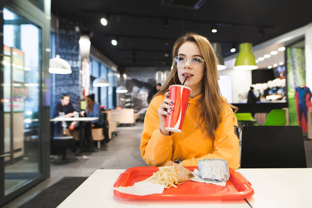 Chica atractiva en vasos sentados en restaurante de comida rápida con una bandeja de papas fritas y hamburguesa y beber cola de la taza. Estudiante chica se sienta en un restaurante y come comida rápida
. - Foto, imagen