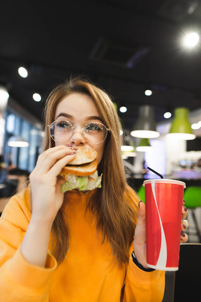 Portré egy vicces szép lány, aki eszik egy finom hamburgert, és egy pohár ital tartja a kezében. A vonzó lány eszik gyorsételek, könnyű gyorsétteremben. Lány eszik egy hamburgert - Fotó, kép