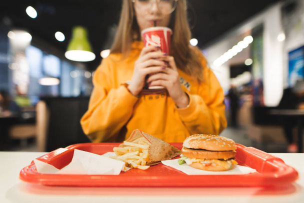 Vassoio dal menu del fast food: patatine fritte, hamburger sullo sfondo intelligente della ragazza che beve. La ragazza in abiti arancioni mangia fast food al ristorante. Concentrati sul vassoio con il cibo
. - Foto, immagini