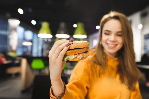 Positive beautiful girl sits in fast food restaurant, holds tasty big burger in hand and smiles. Teen girl eats a burger. Focus on a fresh appetizing burger in the hands of the girl. - Photo, Image