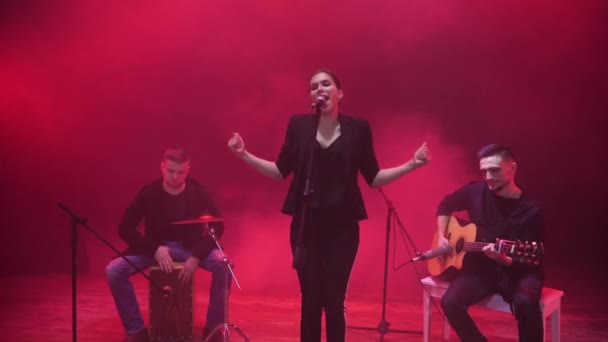 Music band performing on stage in red light and smoke in background. Three musicians - girl singer, drummer and guitarist perform music during the show. - Footage, Video
